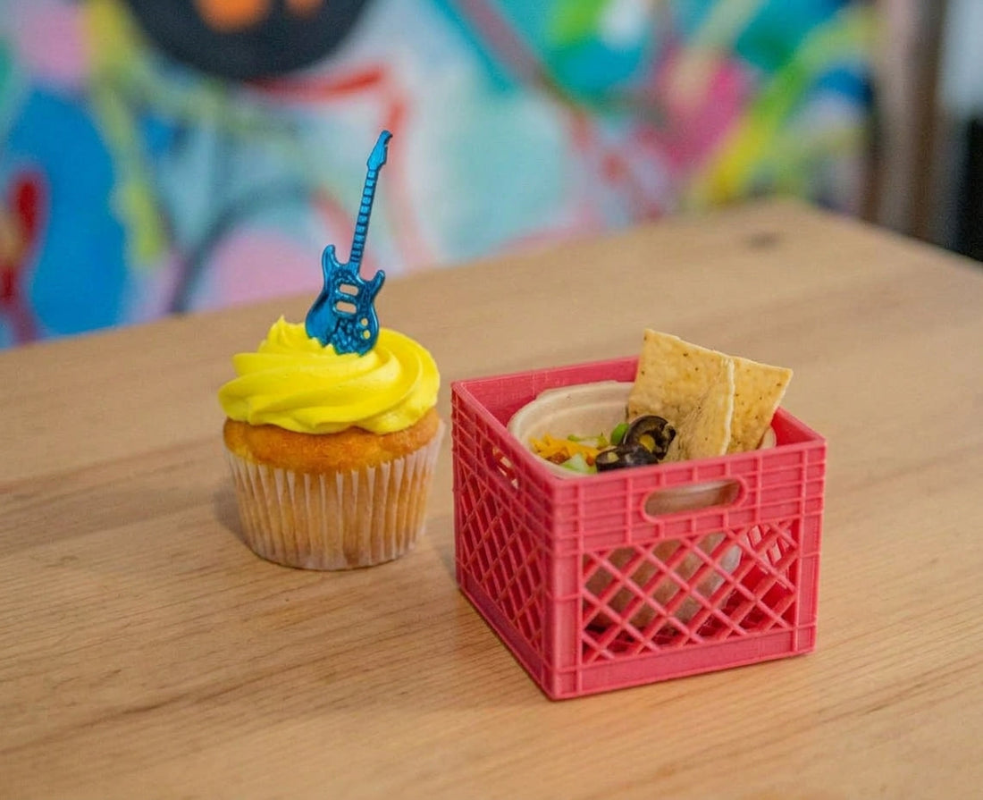Yellow frosted cupcake topped with a blue guitar shaped topper beside a small red 3D printed milk crate holding chips and dip on a wooden table