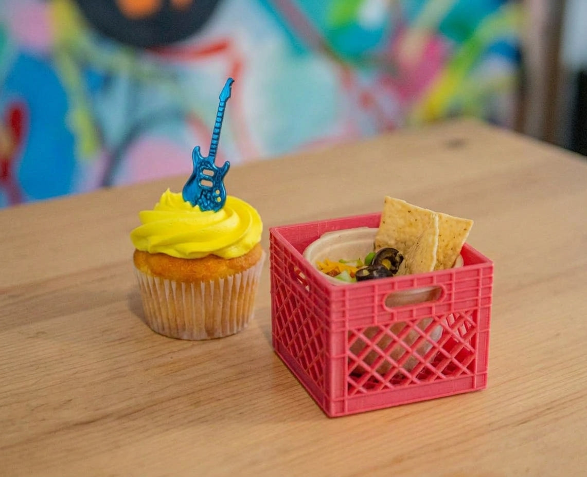 Yellow frosted cupcake topped with a blue guitar shaped topper beside a small red 3D printed milk crate holding chips and dip on a wooden table