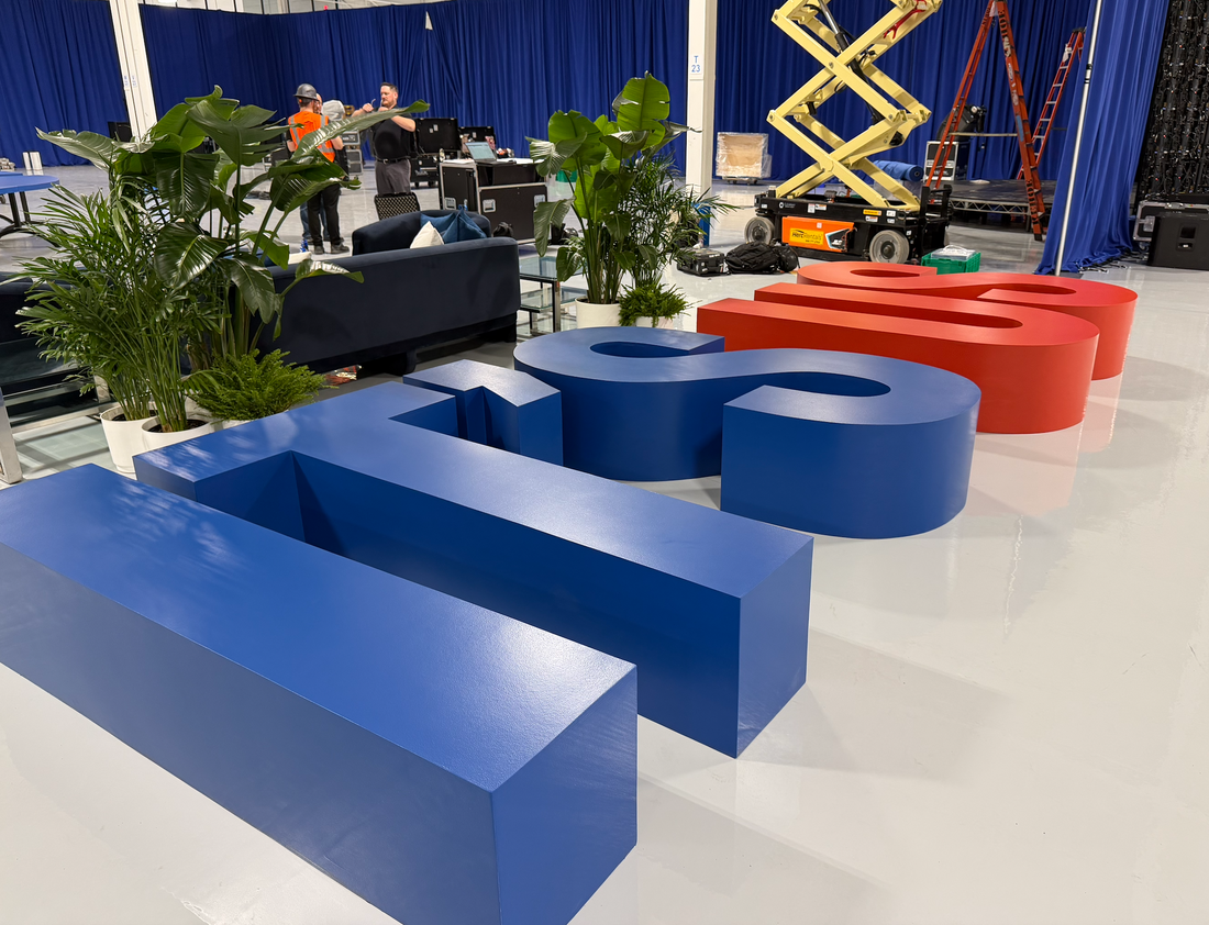 Oversized dimensional letter benches in blue and red with a smooth painted finish, set up on a white floor during an event installation with plants and lounge seating nearby