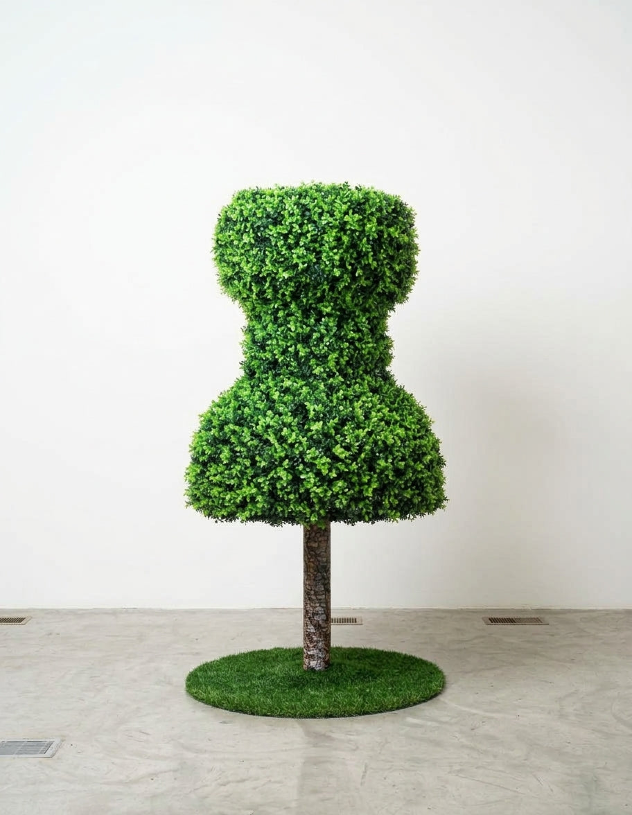 Seven foot tall topiary sculpture shaped like a giant push pin, covered in faux boxwood greenery on a bark textured trunk, standing on a round artificial grass base