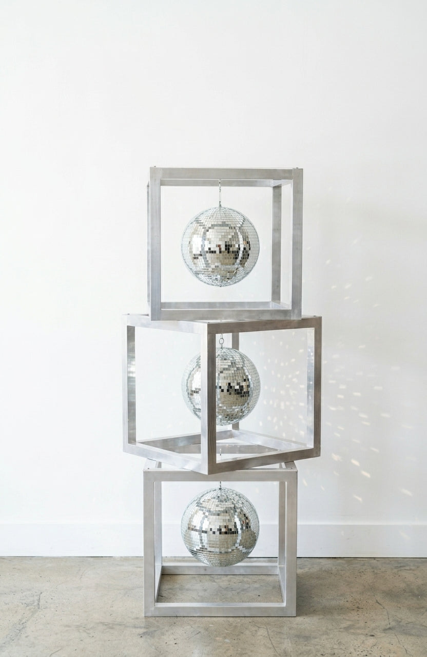 Three stacked open cube frames with a silver metallic finish, each with a hanging mirrored disco ball inside, casting light spots on a white wall