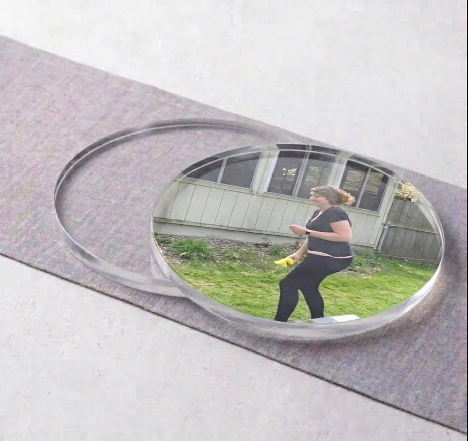 A small oval mirror with a clear frame resting on a gray textured mat against a light gray background. The mirror reflects a woman standing outdoors on grass in front of a house.